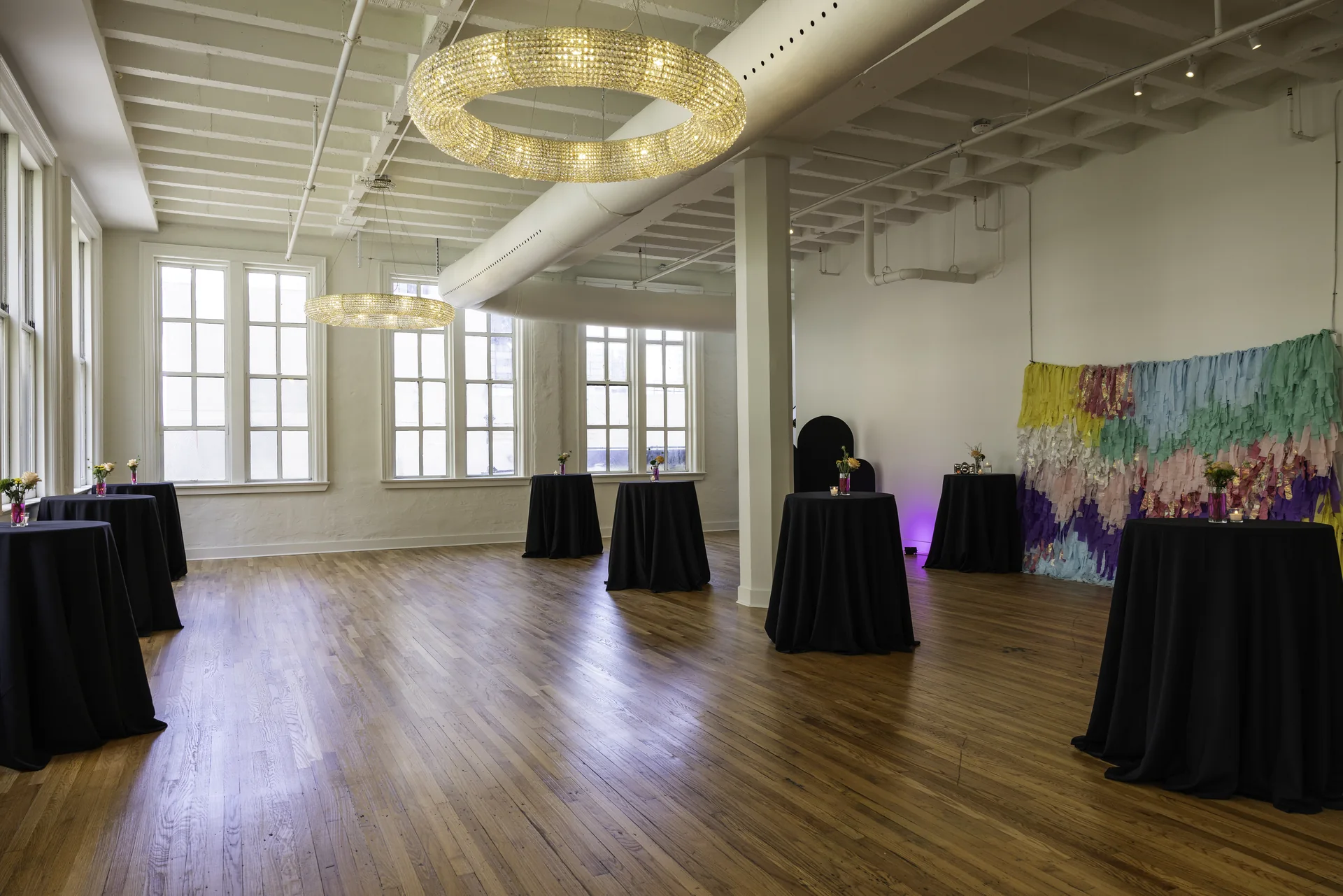 Grand Ballroom’s sunlit corner with tall windows, a crystal chandelier, open wood floor and cocktail tables.