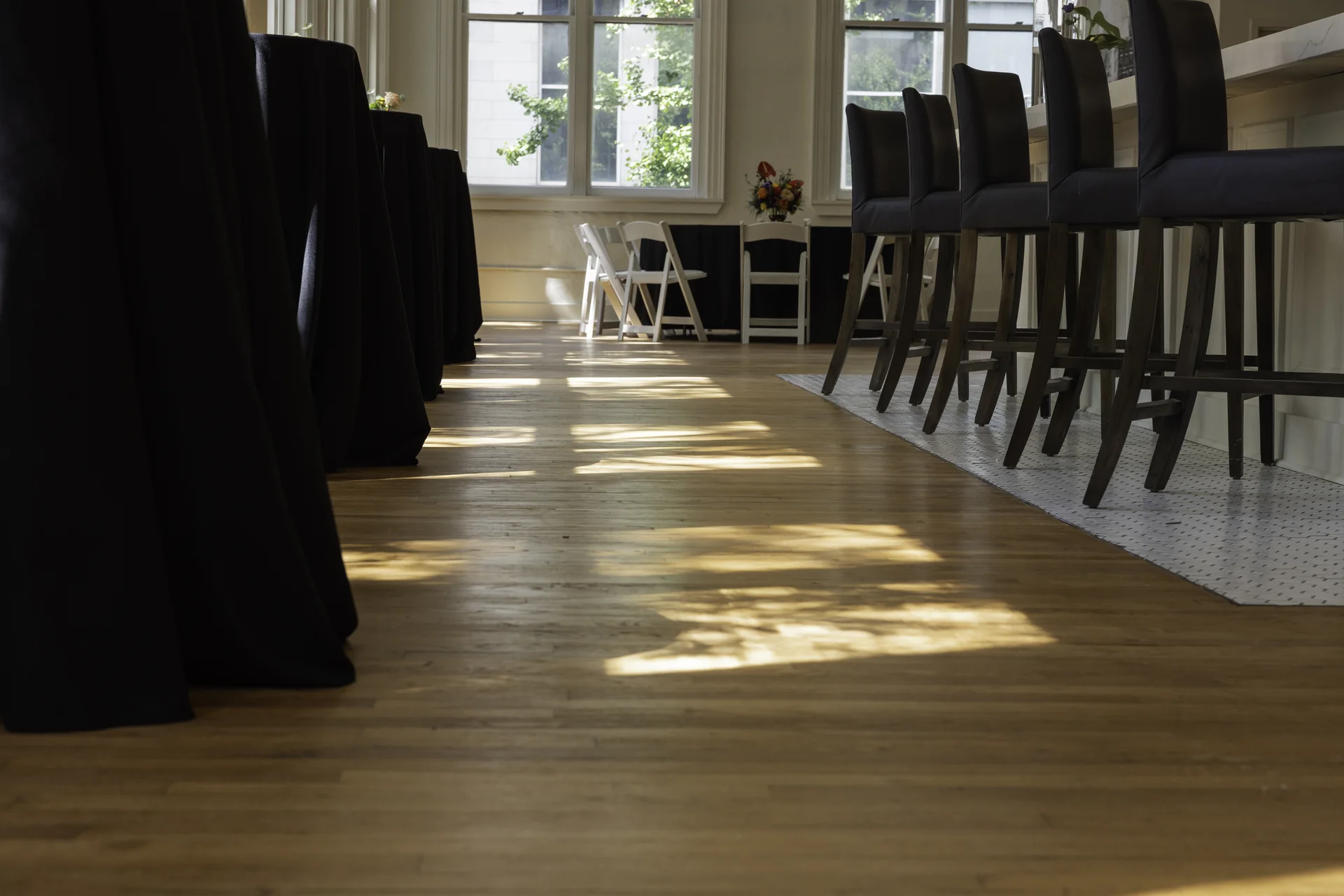 A low view along Grand Ballroom’s hardwood floor with sunlight, bar stools and tall windows.